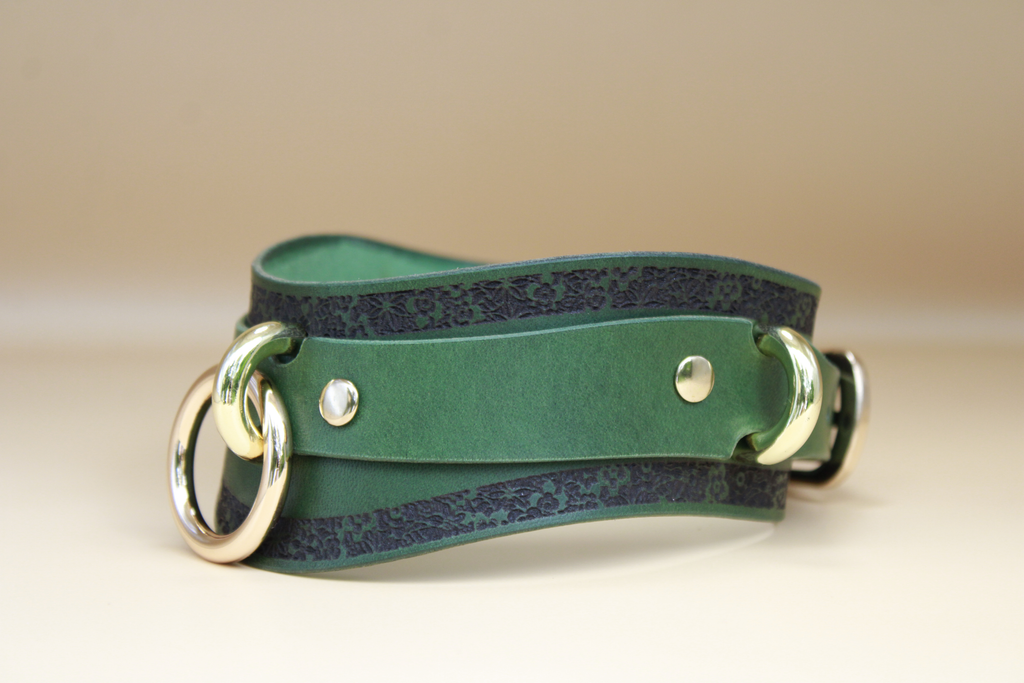 A green leather choker with a dark green floral pattern and two gold-colored metal rings on either side.