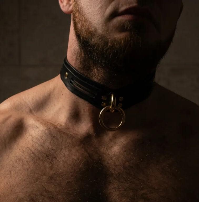 A shirtless man with a beard wearing a black leather choker necklace with a gold ring.