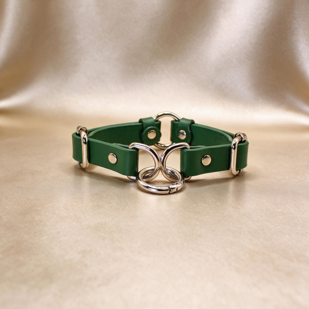 A green leather choker necklace with metal rings and buckles is displayed on a beige background.