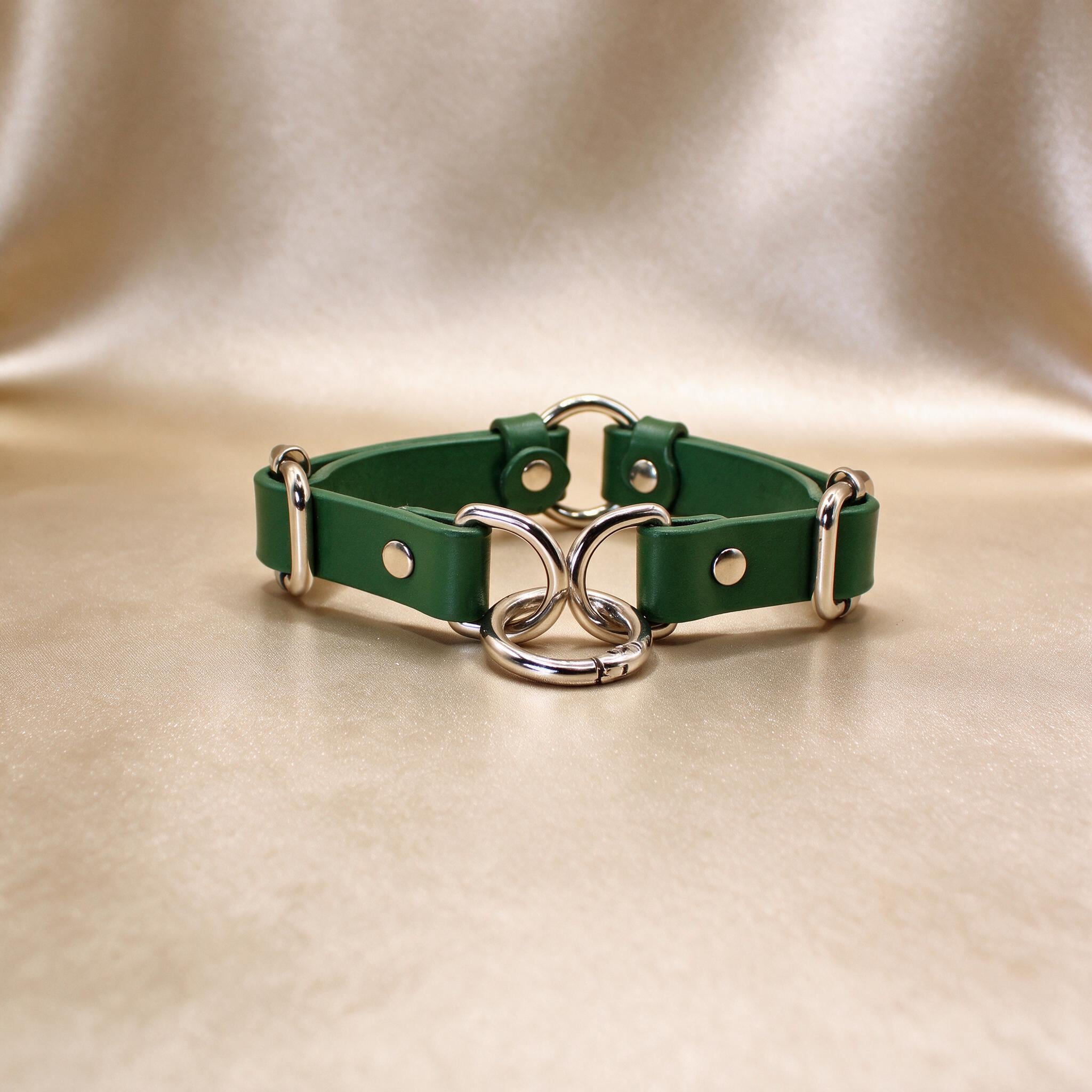 A green leather choker necklace with metal rings and buckles is displayed on a beige background.