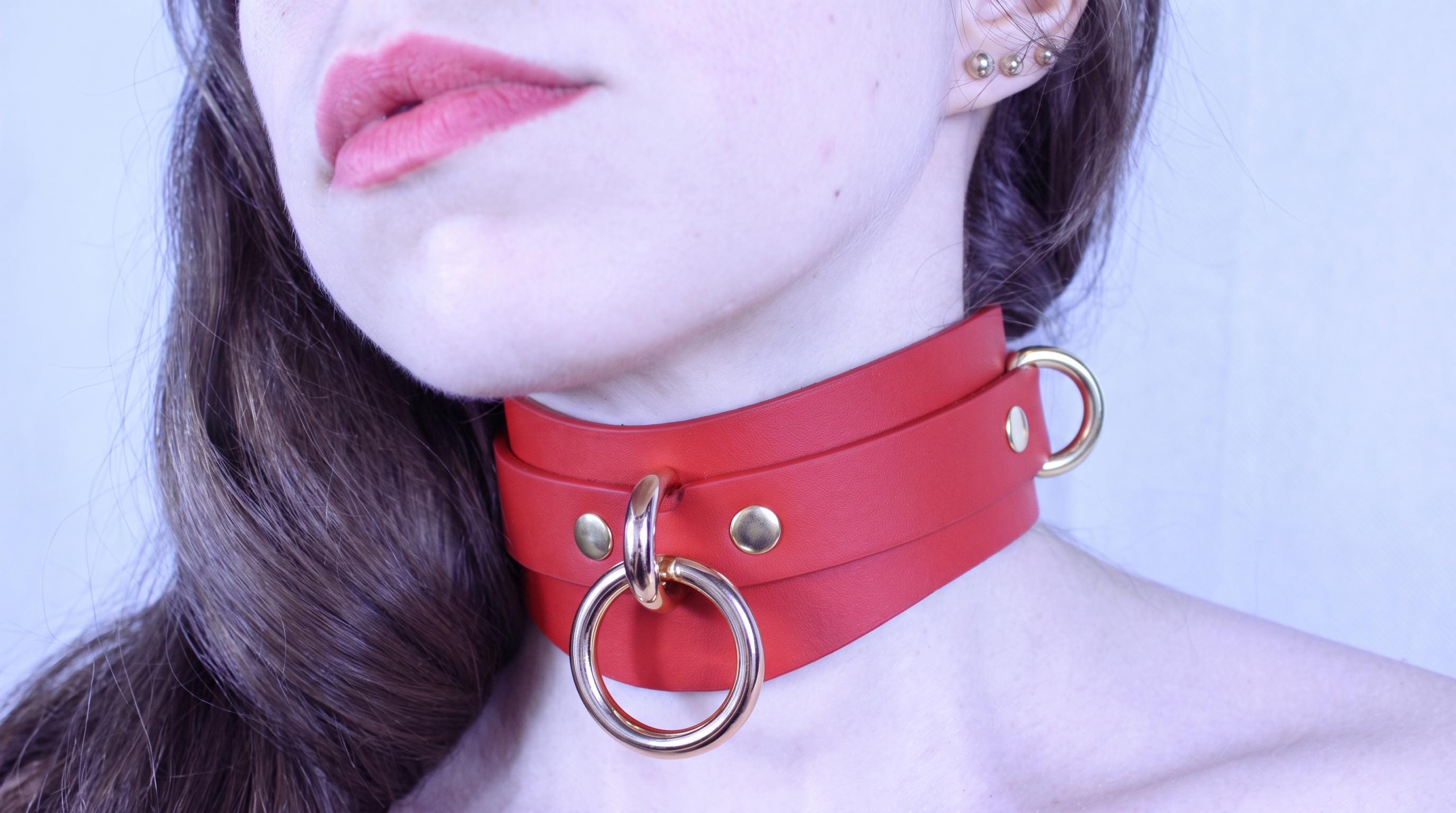 A close-up of a woman's face wearing a red leather choker necklace with gold hardware.