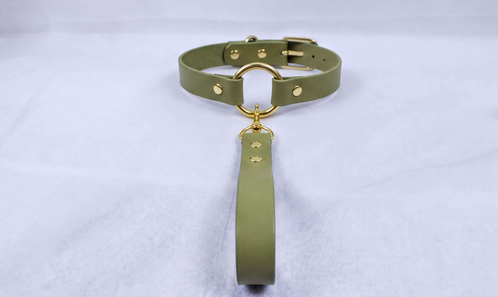 A green choker collar with a gold-colored metal ring and buckle is displayed on a white surface.