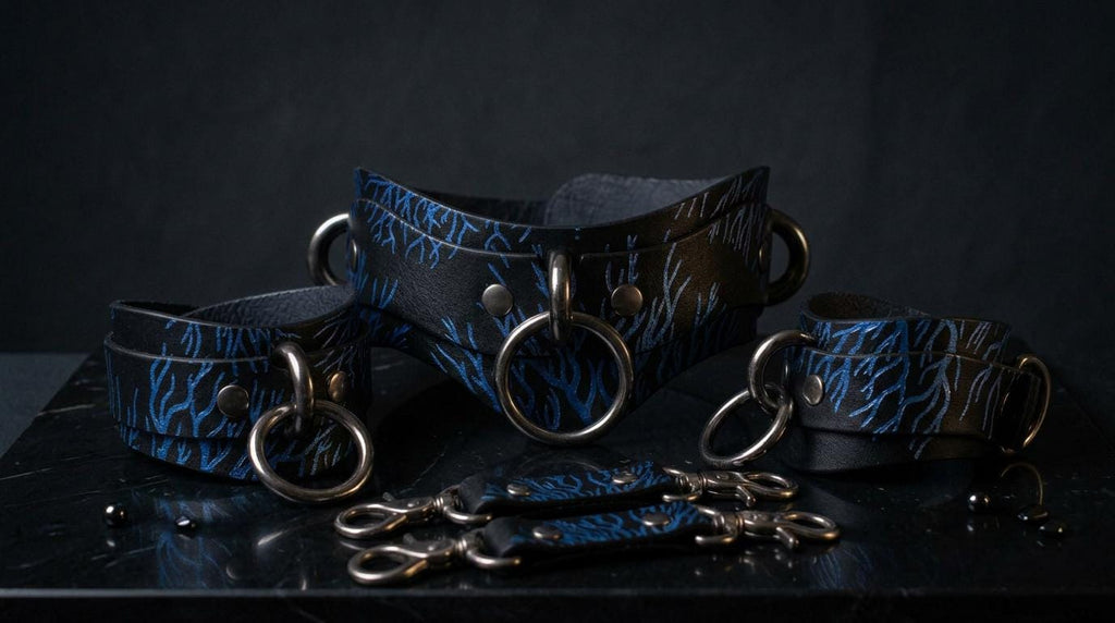 A set of black leather cuffs with blue floral patterns, featuring metal rings and buckles, displayed on a dark surface.