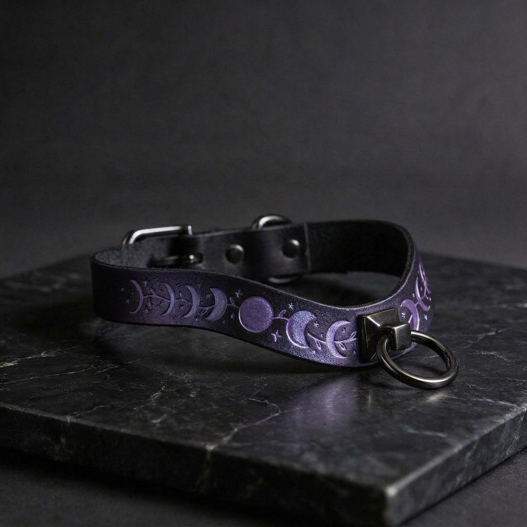 A black leather collar with a purple moon and star pattern on it, featuring a metal ring and buckle.