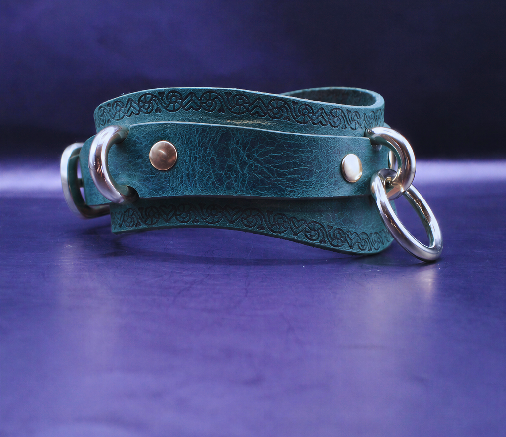 A teal-colored leather choker necklace with metal rings and a decorative pattern on the clasp is displayed against a dark purple background.