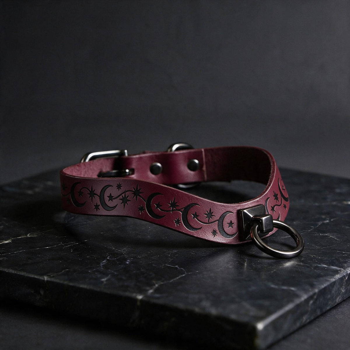 A burgundy leather collar with a star and crescent design, featuring a metal ring and buckle, is displayed on a dark surface.