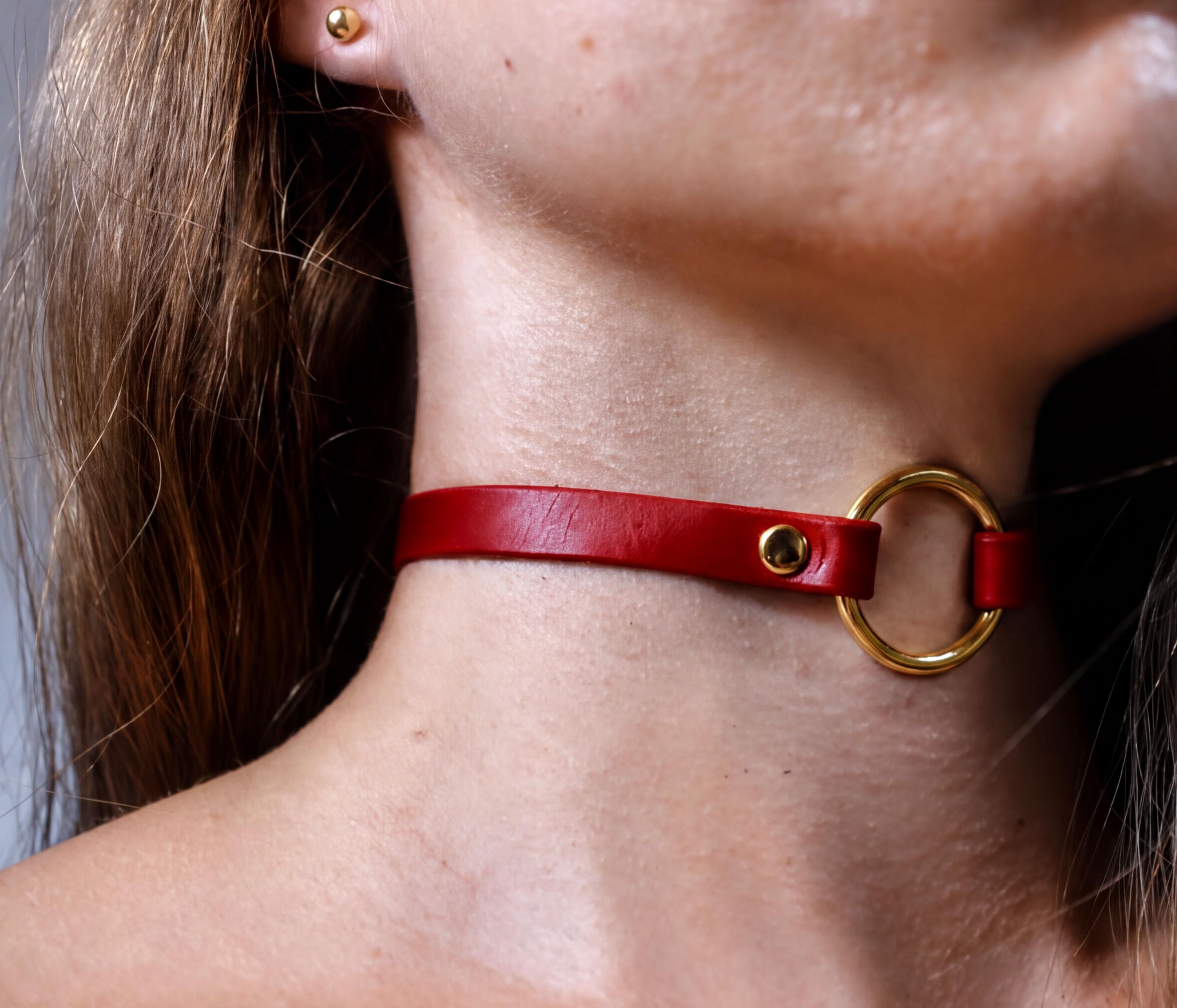 A close-up of a person's neck and chin wearing a red leather choker necklace with a gold ring.