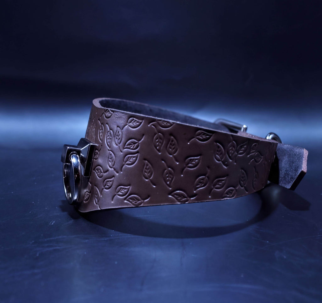 A brown leather belt with a leaf pattern design, featuring a metal buckle and a dark background.