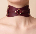 A close-up view of a burgundy leather choker necklace with a gold ring and two metal studs on the front.
