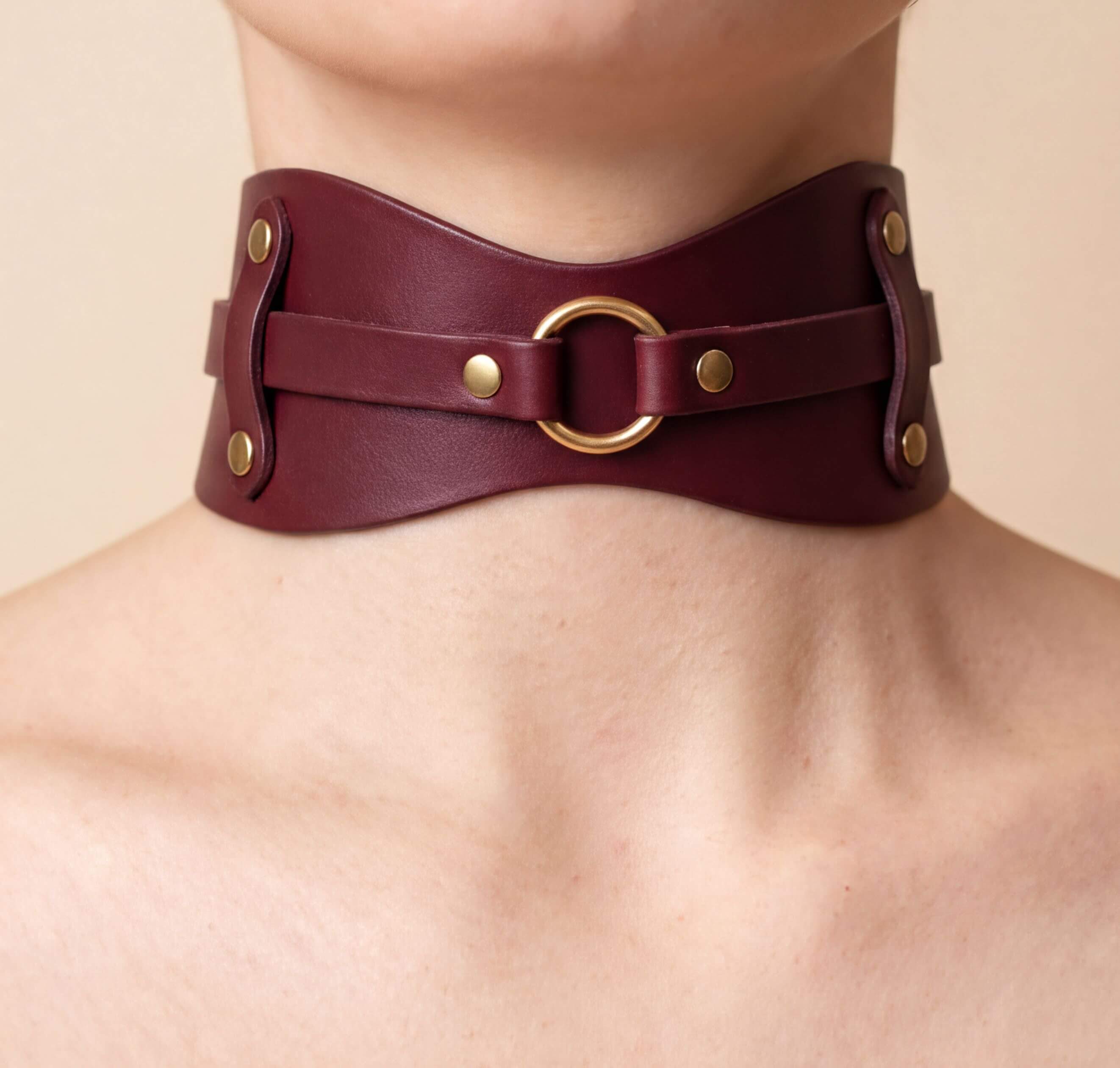 A close-up view of a burgundy leather choker necklace with a gold ring and two metal studs on the front.