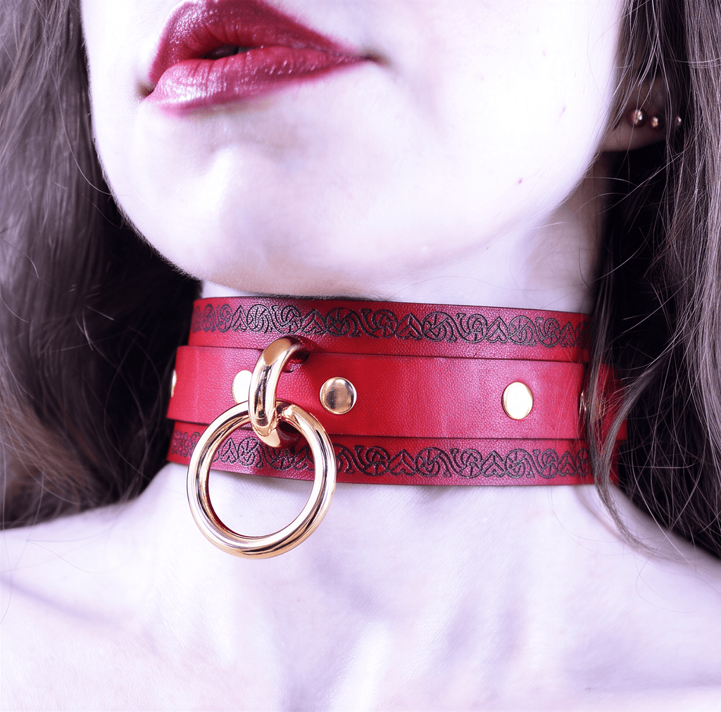 A close-up of a woman's neck and chest area wearing a red leather choker necklace with a gold ring.