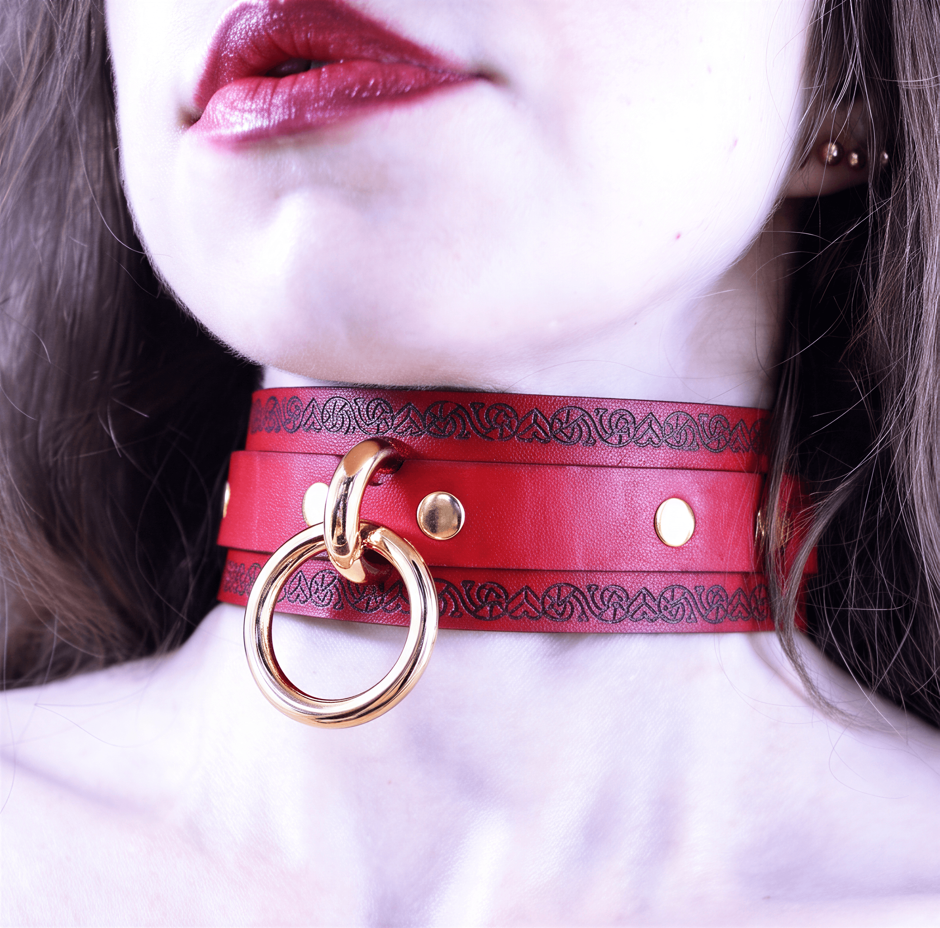 A close-up of a woman's neck and chest area wearing a red leather choker necklace with a gold ring.