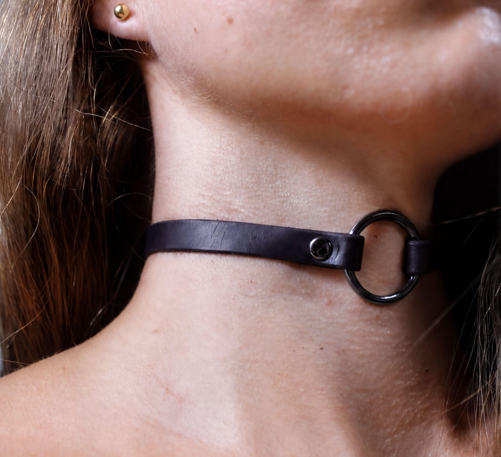 A close-up view of a person's neck and chin area, featuring a black leather choker necklace with a metal ring.