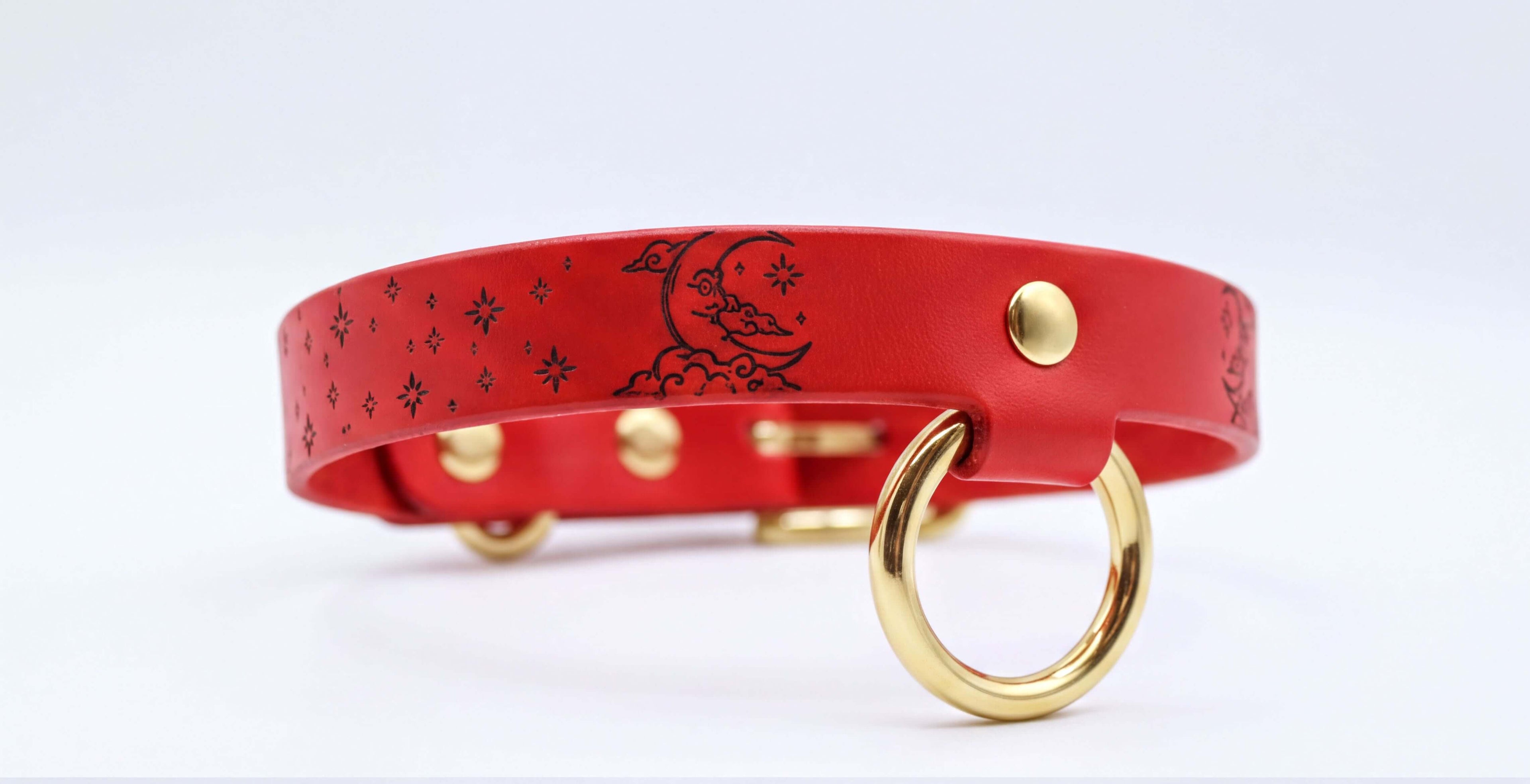 A red leather collar with a moon and star design, featuring a gold ring and buckle.