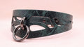 A close-up view of a dark green leather choker necklace with a metal ring and cutout design.