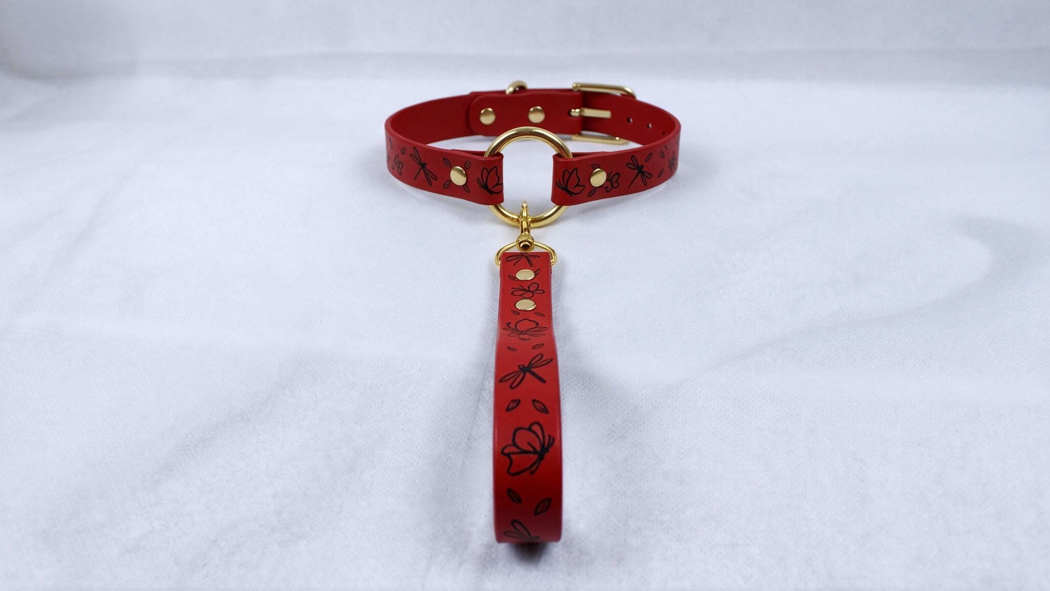 A red collar with a gold buckle and a red leash attached to it, placed on a white surface.