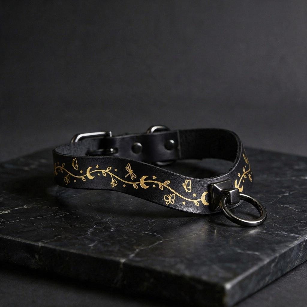A black leather collar with a gold floral design and a metal ring on the front, resting on a dark surface.