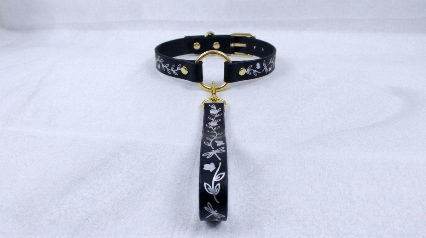 A black collar with a gold-colored buckle and a floral pattern on the strap is displayed against a white background.