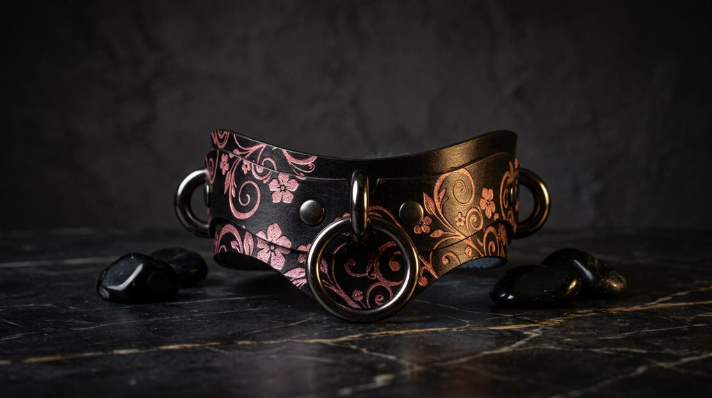 A decorative leather collar with floral patterns and metal rings is displayed on a dark surface, accompanied by small black stones.