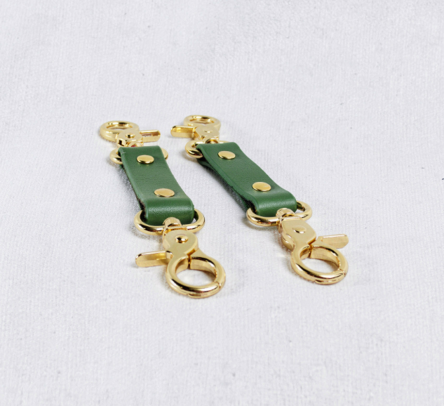 A pair of green leather straps with gold metal clasps and hooks, arranged side by side on a white surface.