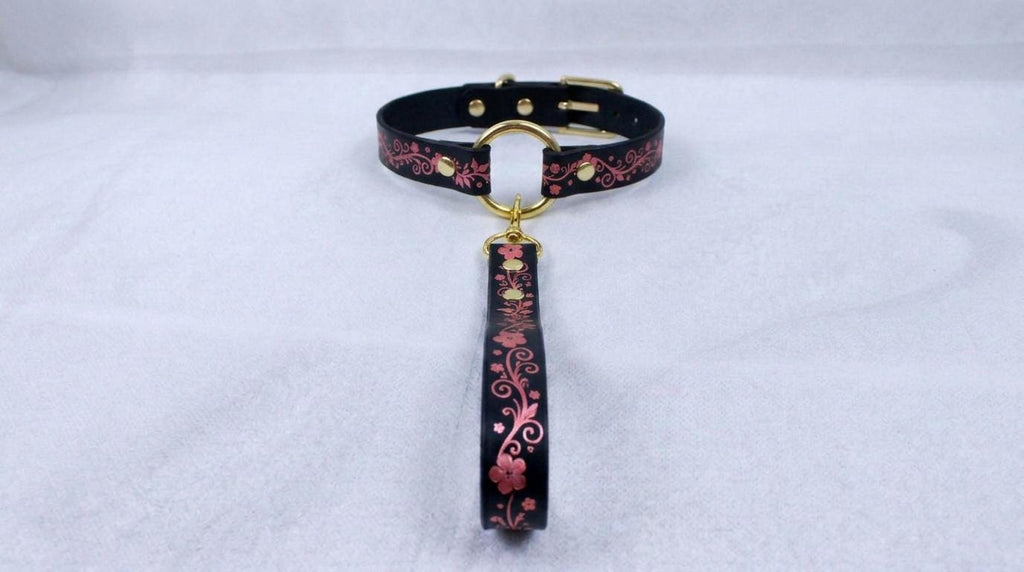 A black collar with a floral pattern and a gold-colored buckle is displayed on a white surface.