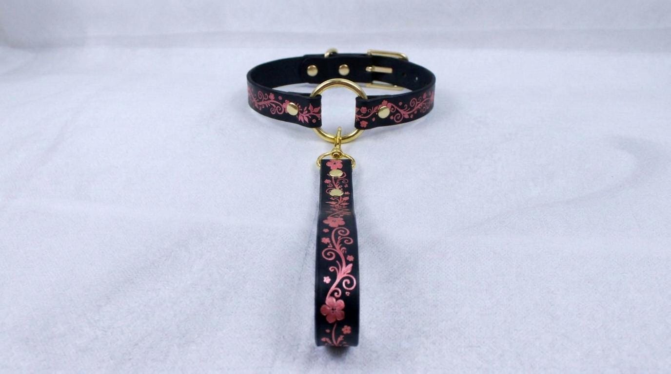 A black collar with a floral pattern and a gold-colored buckle is displayed on a white surface.