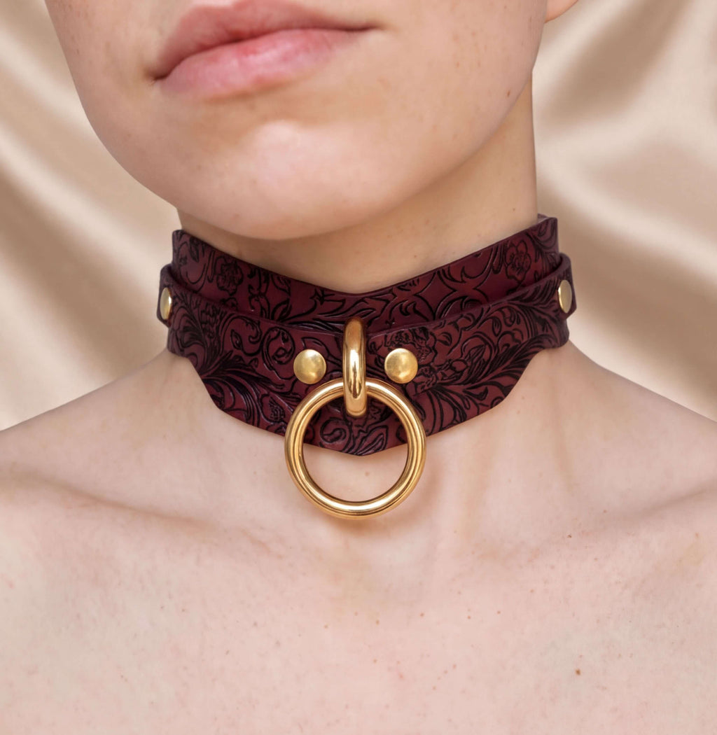 A close-up of a person's neck wearing a burgundy choker necklace with a gold ring and buckle.