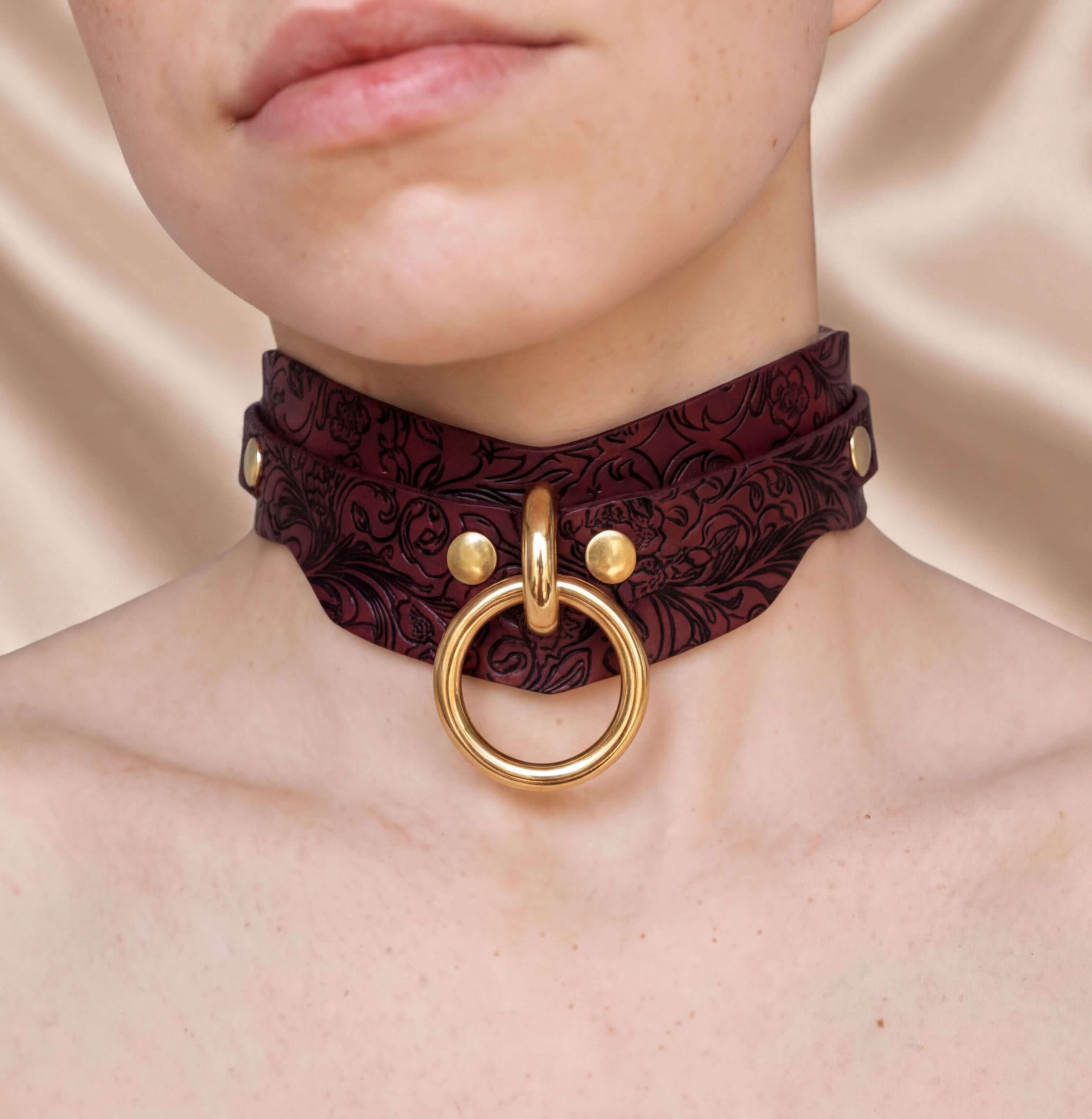 A close-up of a person's neck wearing a burgundy choker necklace with a gold ring and buckle.
