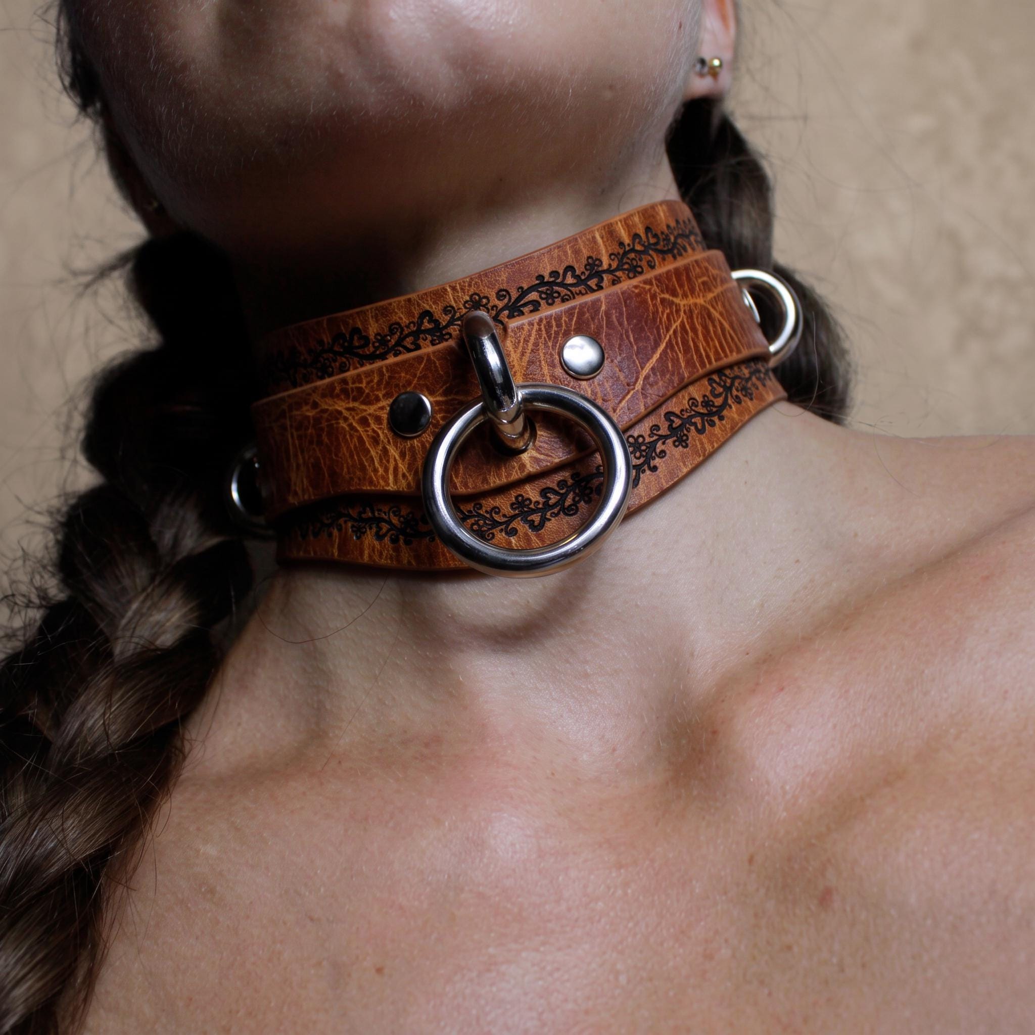 A close-up of a woman's neck wearing a brown leather choker necklace with metal rings and a metal ring.