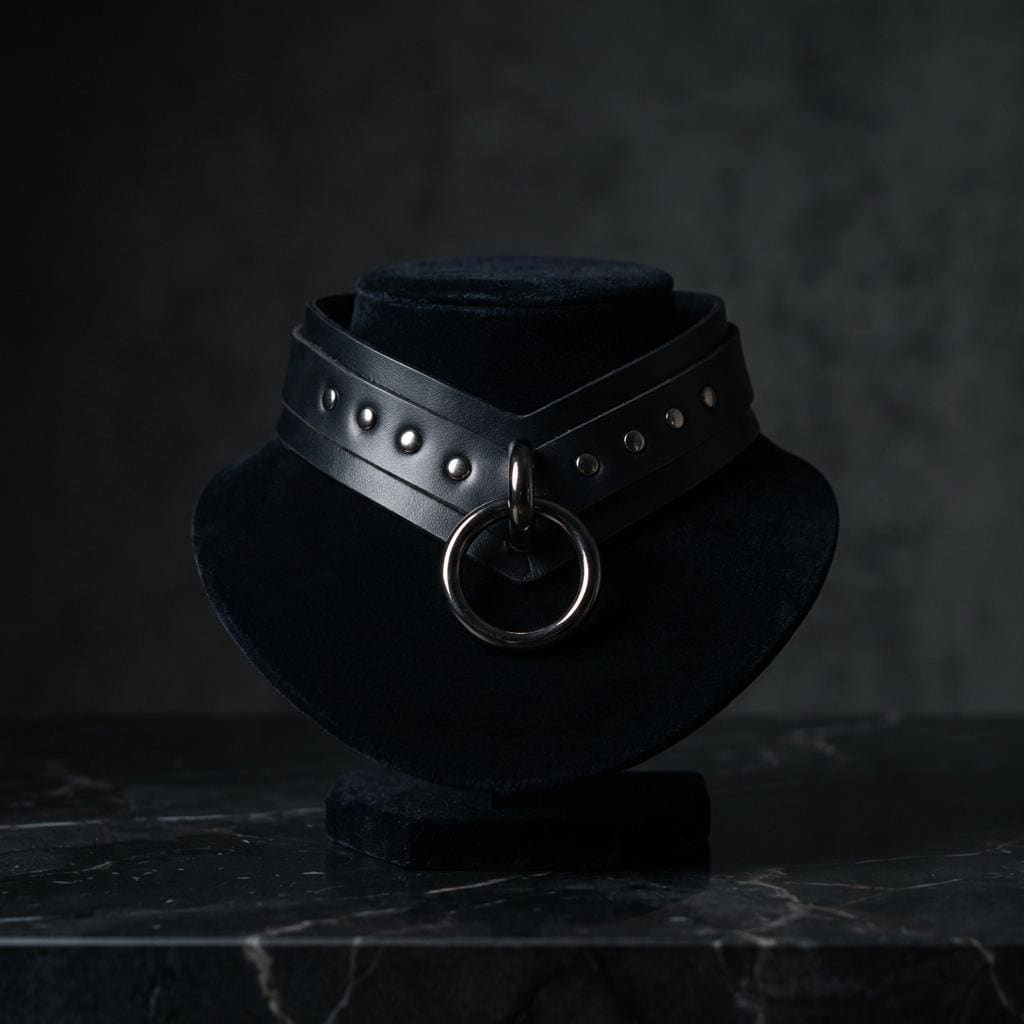 A black leather choker necklace with a large circular metal ring on the front, displayed on a black velvet stand against a dark background.