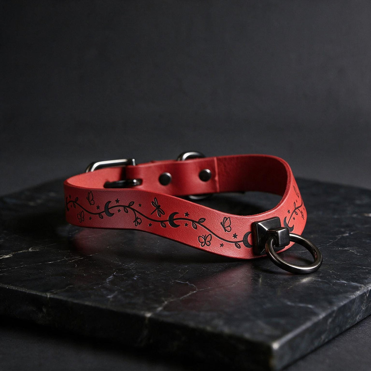 A red leather collar with a metal ring and decorative elements is displayed on a black marble surface.
