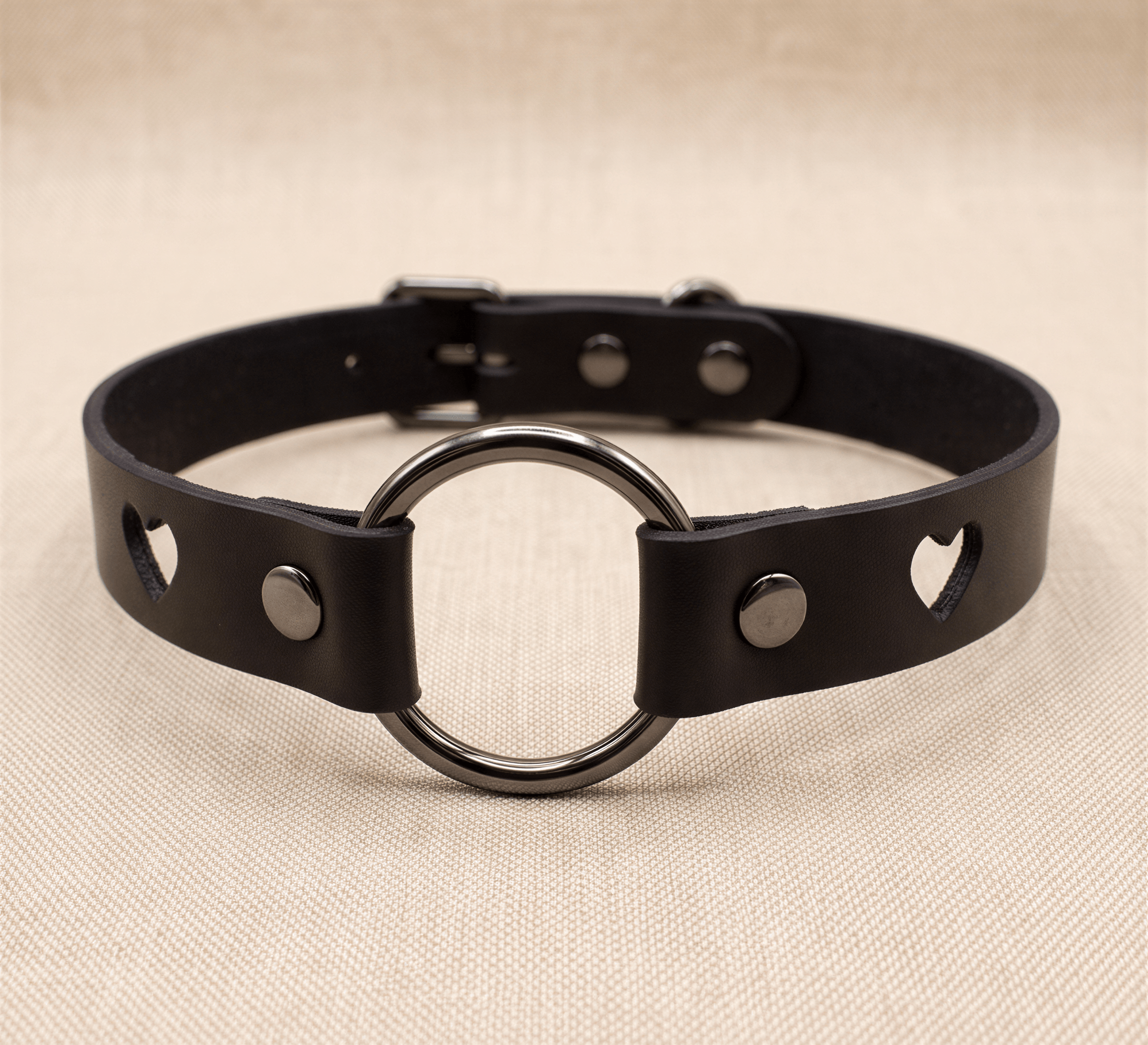 A black leather choker collar with a round metal ring and heart-shaped cutouts on either side.