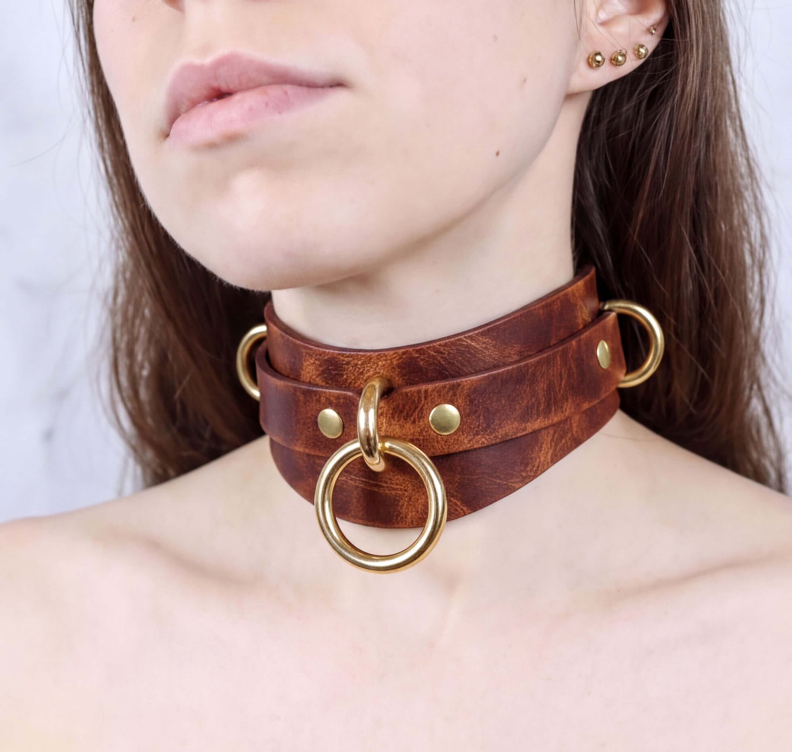 A close-up of a woman's neck wearing a brown leather choker necklace with gold rings and buckles.