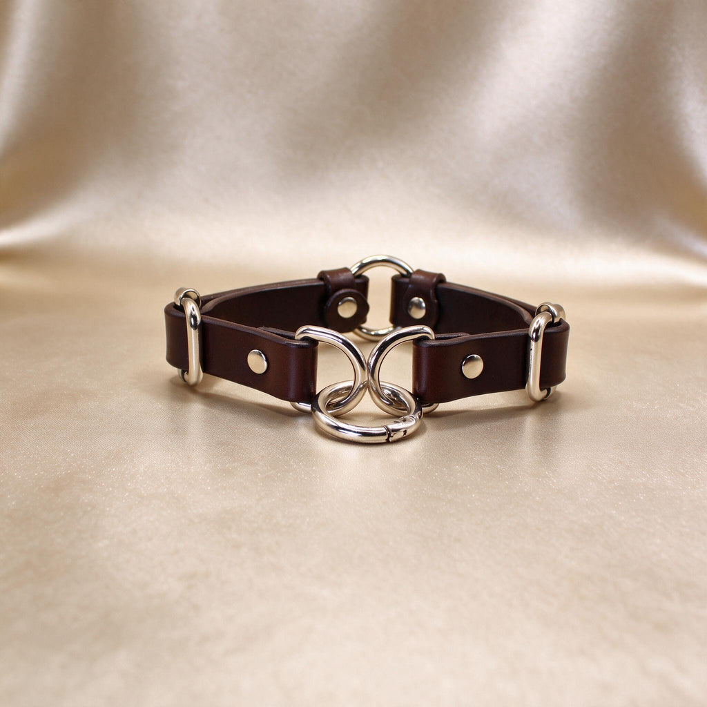 A brown leather choker necklace with silver metal rings and buckles, displayed on a beige background.