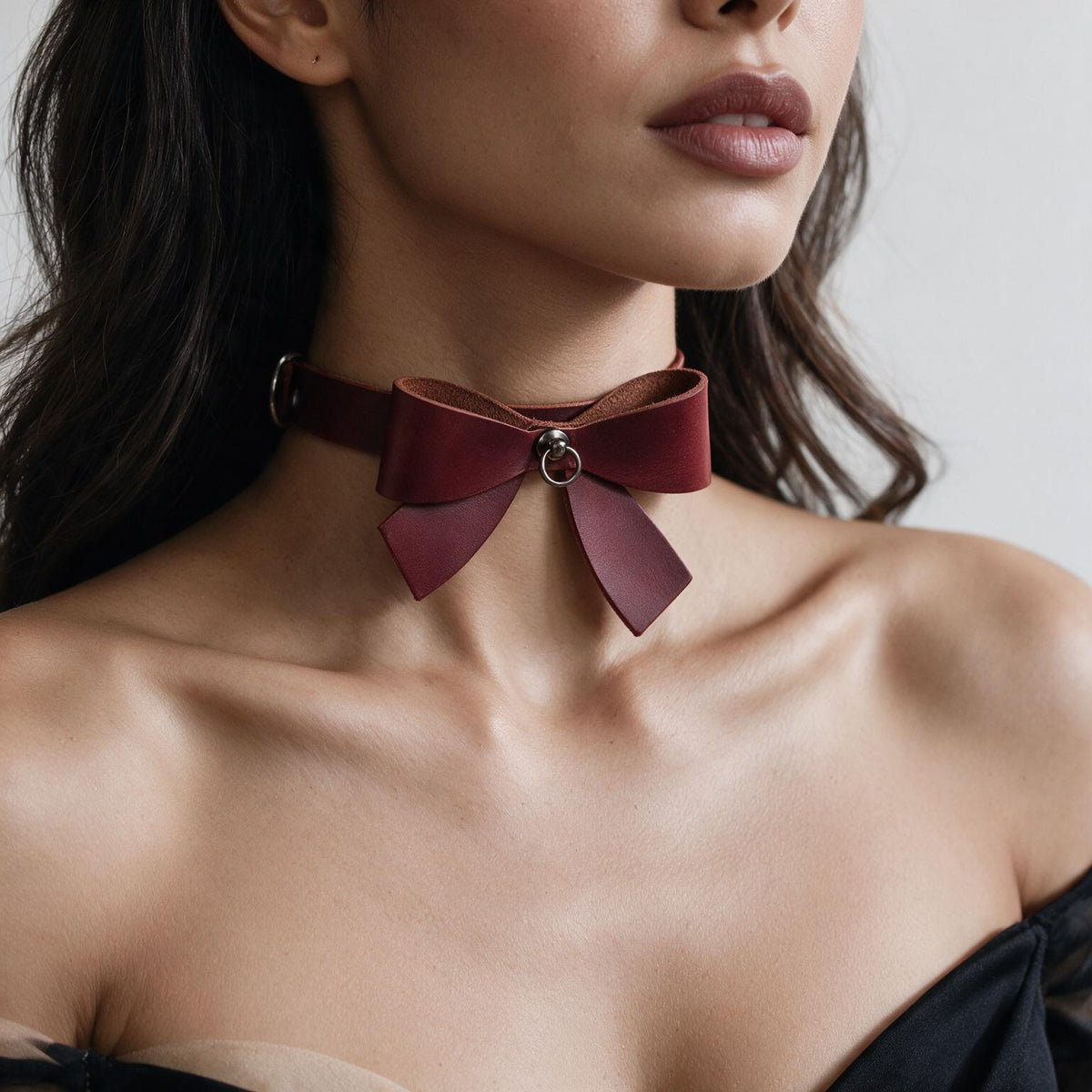 A close-up of a woman's neck and chest area, featuring a burgundy leather choker necklace with a large bow detail.