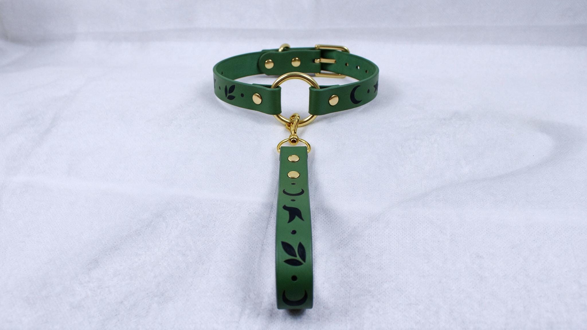 A green dog collar with a gold buckle and a black leaf design on the strap is shown against a white background.