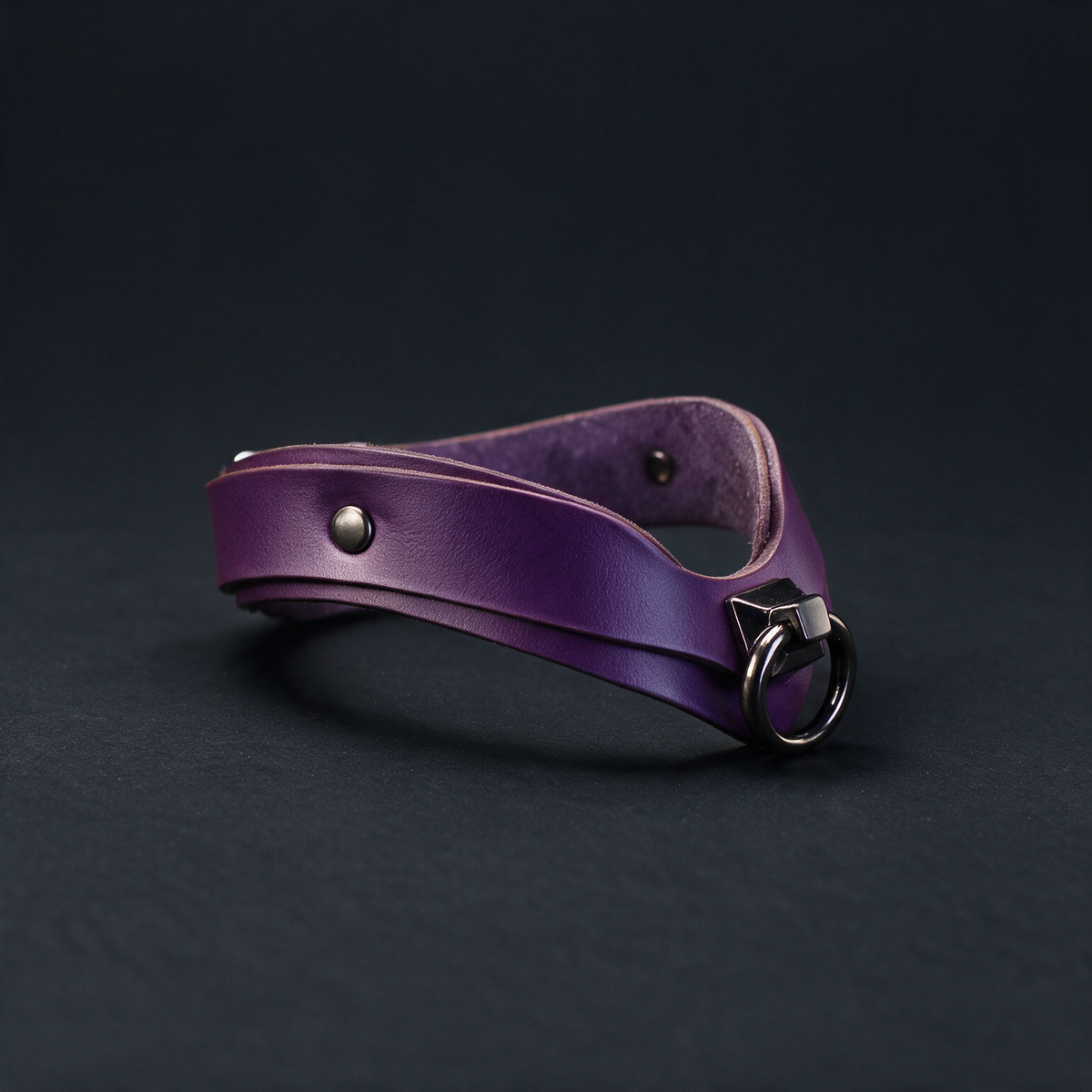 A purple leather choker necklace with a metal clasp and a small metal button on the front is shown against a dark background.