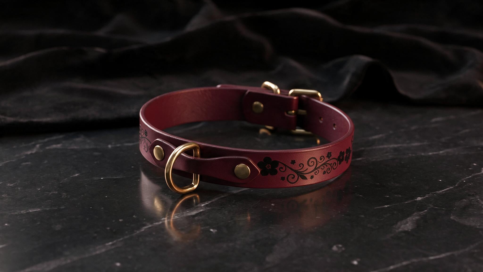 A burgundy leather collar with a decorative pattern and a gold-colored buckle is displayed on a black marble surface.