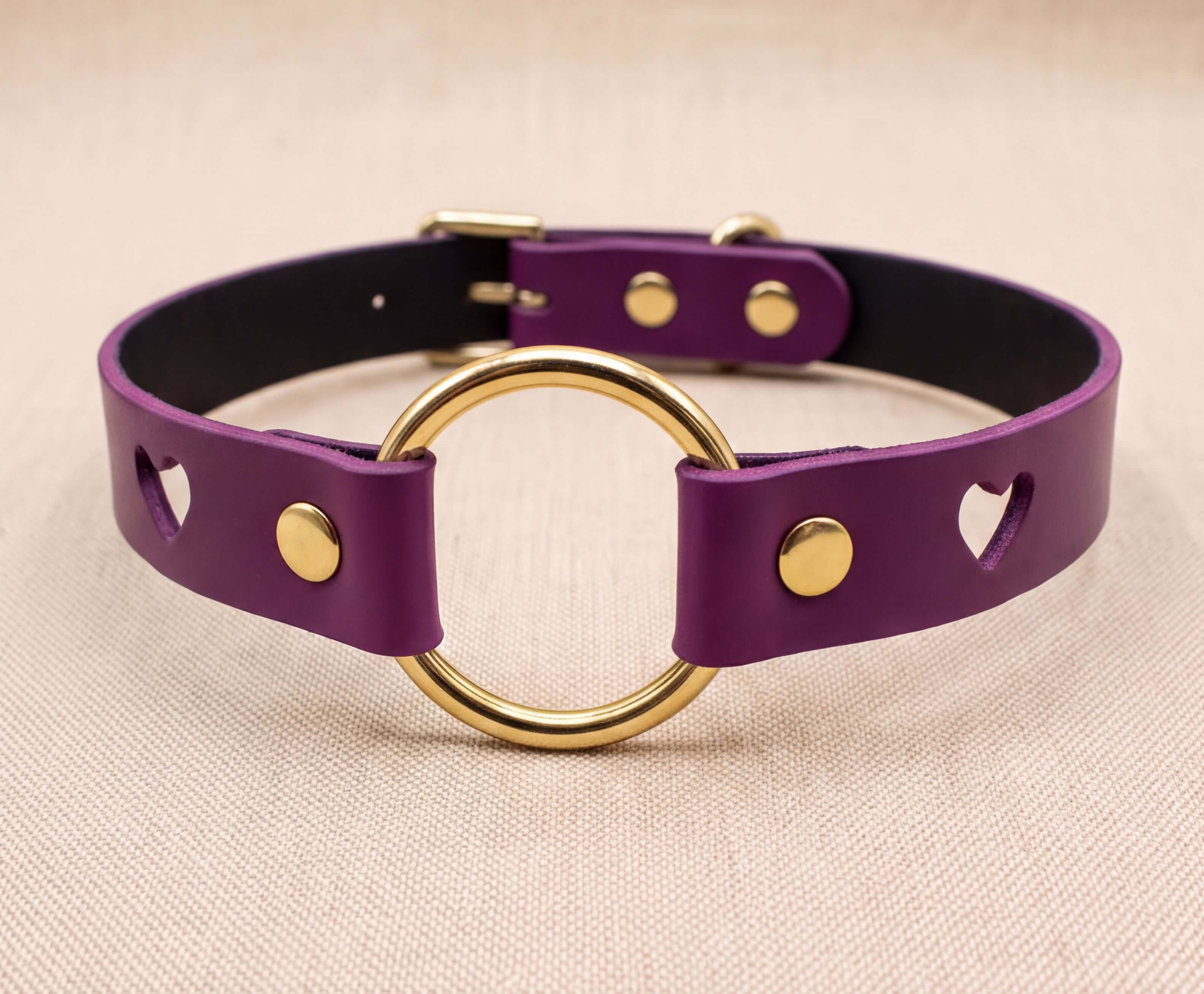 A purple leather choker collar with a gold ring and heart-shaped cutouts on the sides.