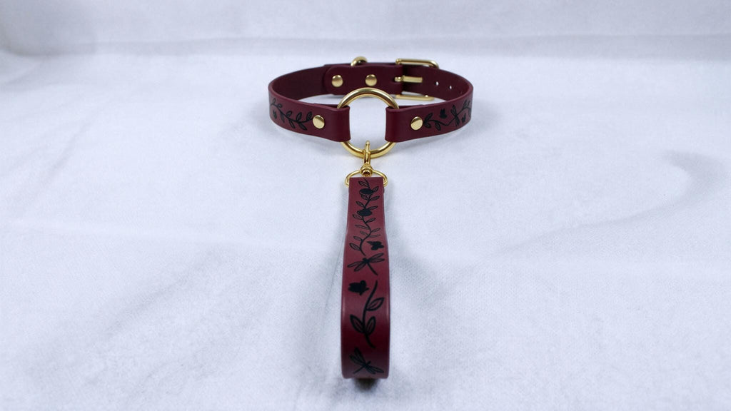 A burgundy leather collar with a gold-colored buckle and a black floral pattern on the strap.