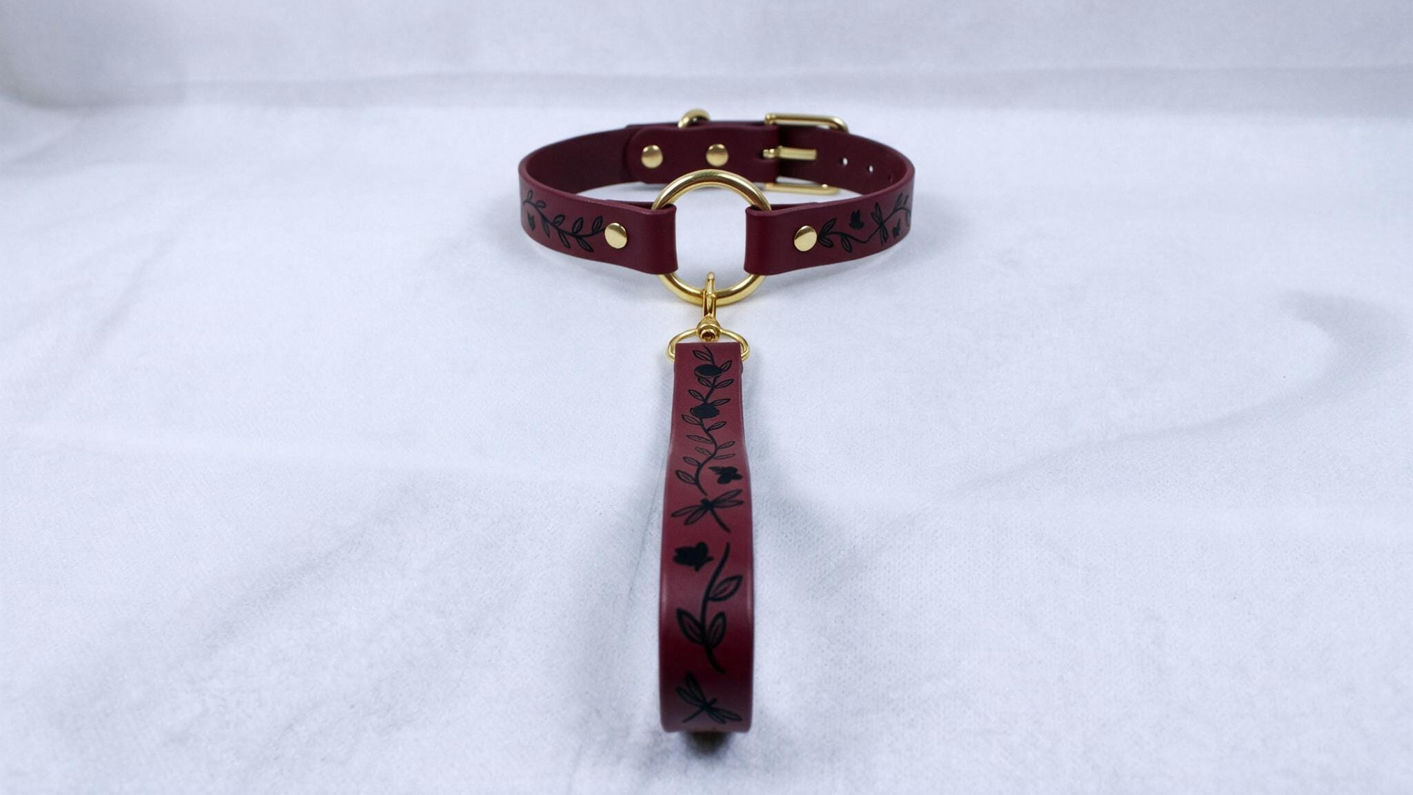 A burgundy leather collar with a gold-colored buckle and a black floral pattern on the strap.