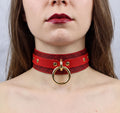 A close-up of a woman's neck and chest area wearing a red choker necklace with a gold ring.