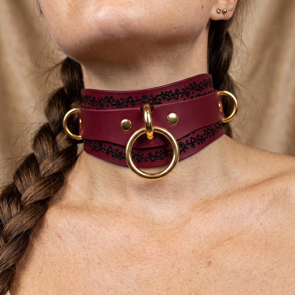 A close-up of a woman's neck and chest area wearing a burgundy leather choker necklace with gold-colored rings and a gold-colored ring on the front.