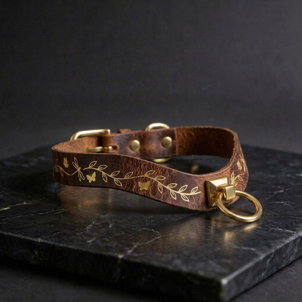 A brown leather collar with gold leaf and butterfly designs, featuring a gold-colored metal ring and buckle, is displayed on a black marble surface.