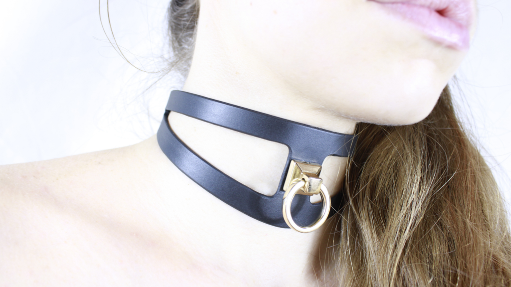 A close-up of a woman's neck and chin wearing a black choker necklace with a gold-colored metal ring.