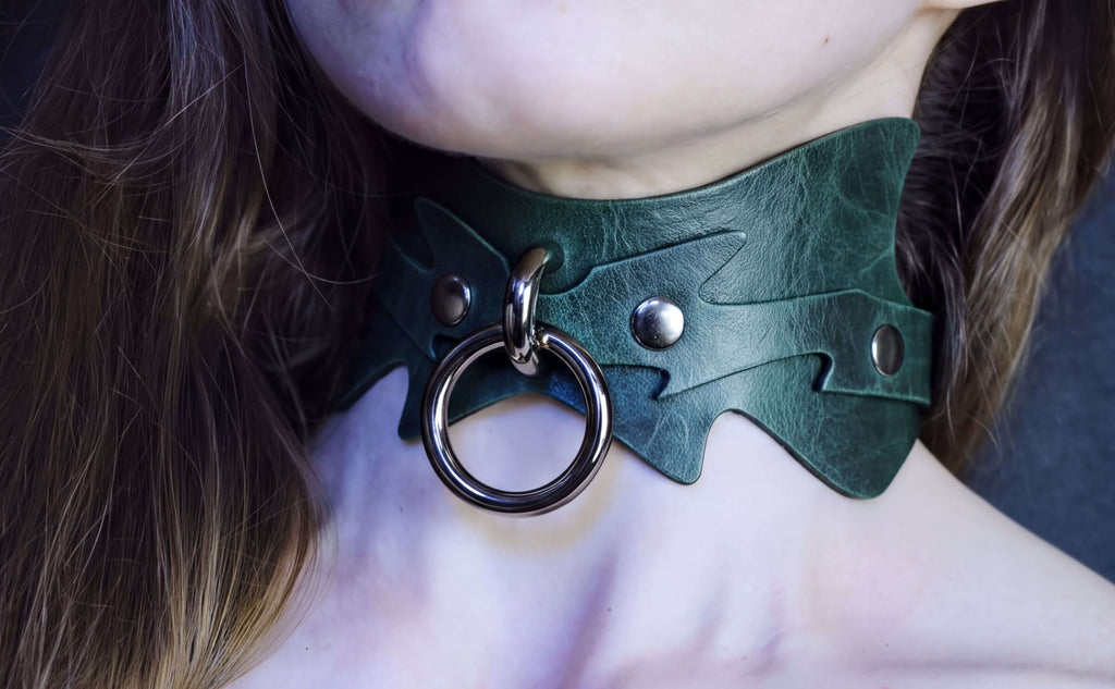 A close-up of a person's neck wearing a green leather choker necklace with a metal ring and two metal studs.