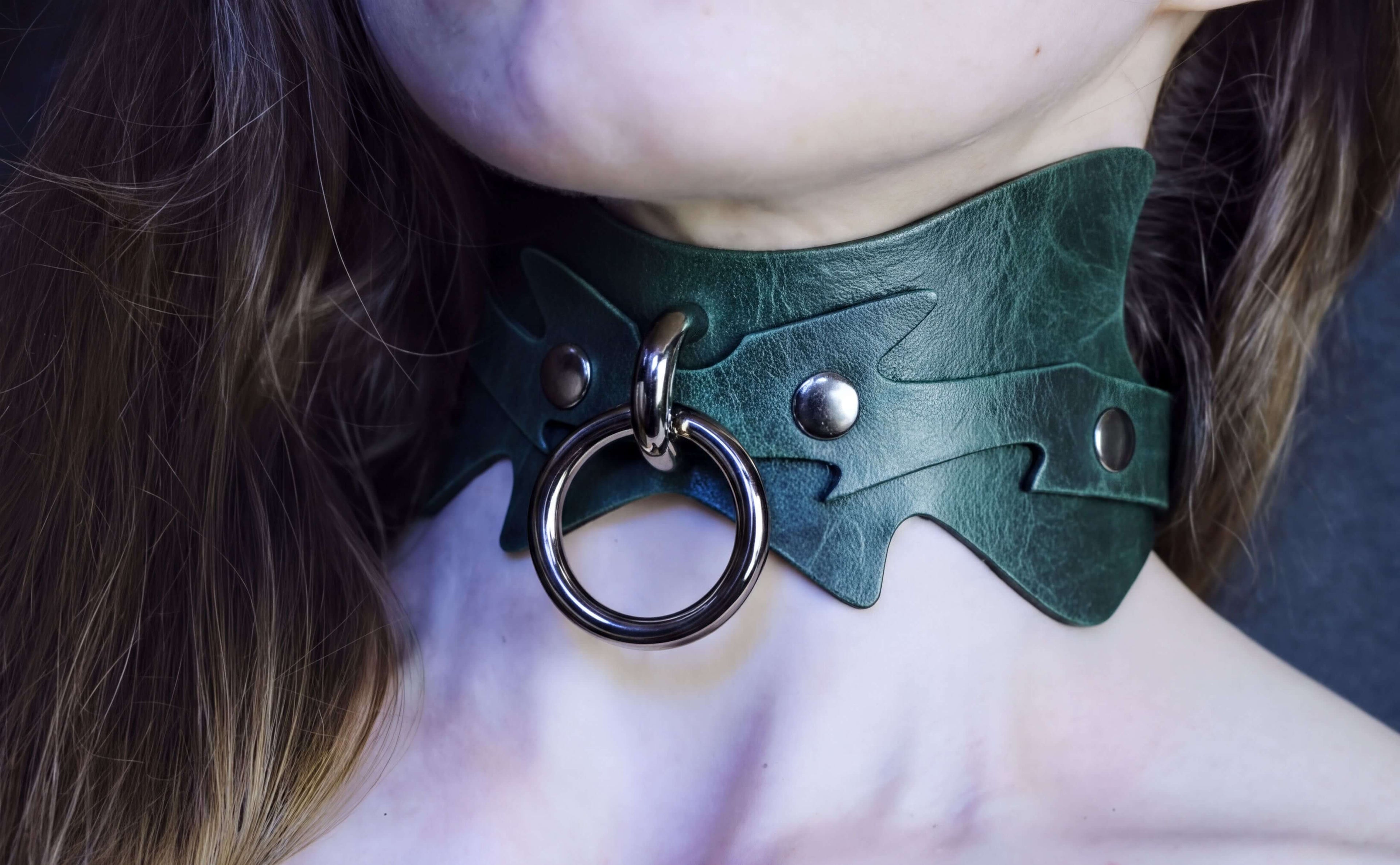 A close-up of a person's neck wearing a green leather choker necklace with a metal ring and two metal studs.