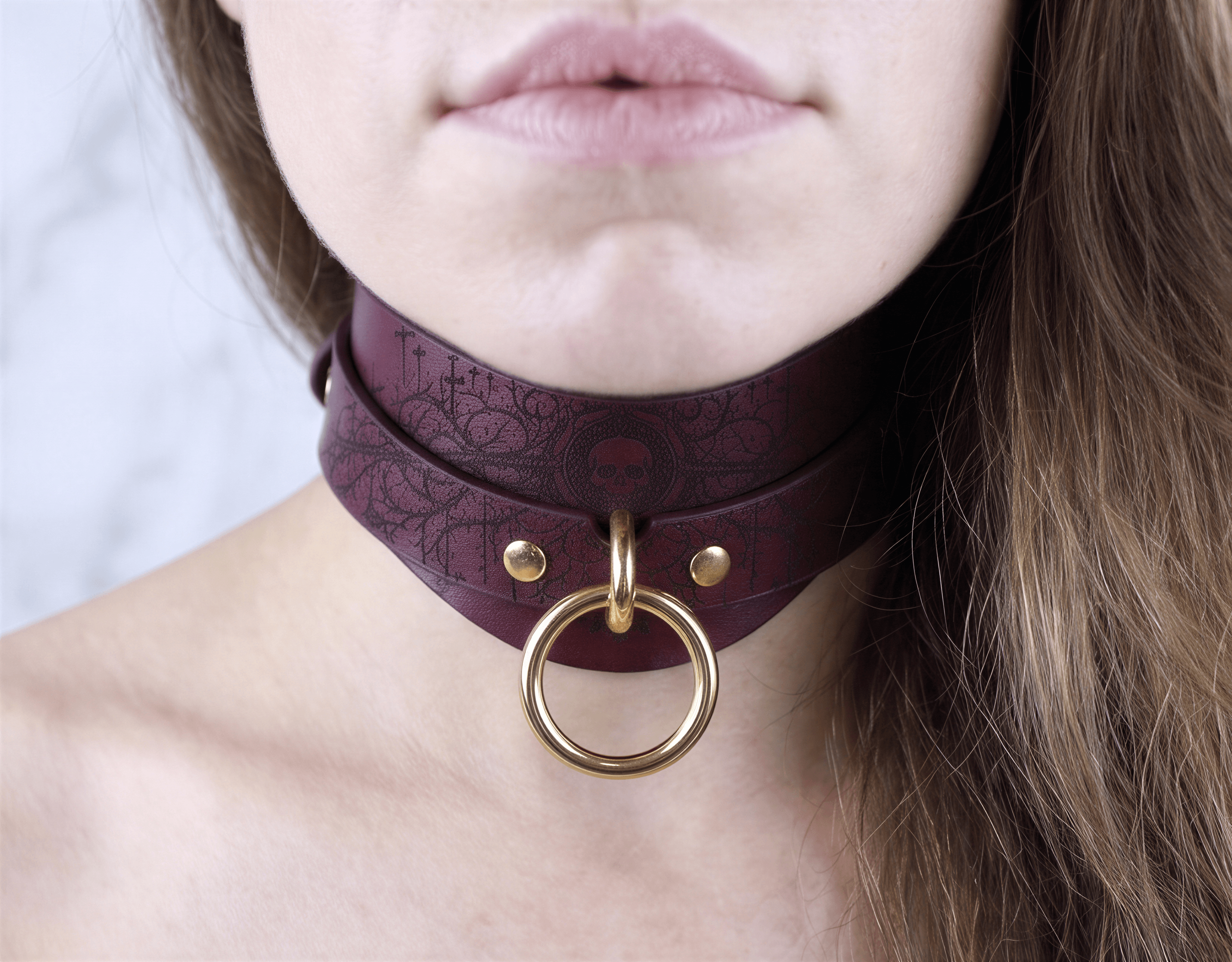 A close-up of a woman's neck and chin wearing a burgundy choker necklace with a gold ring.