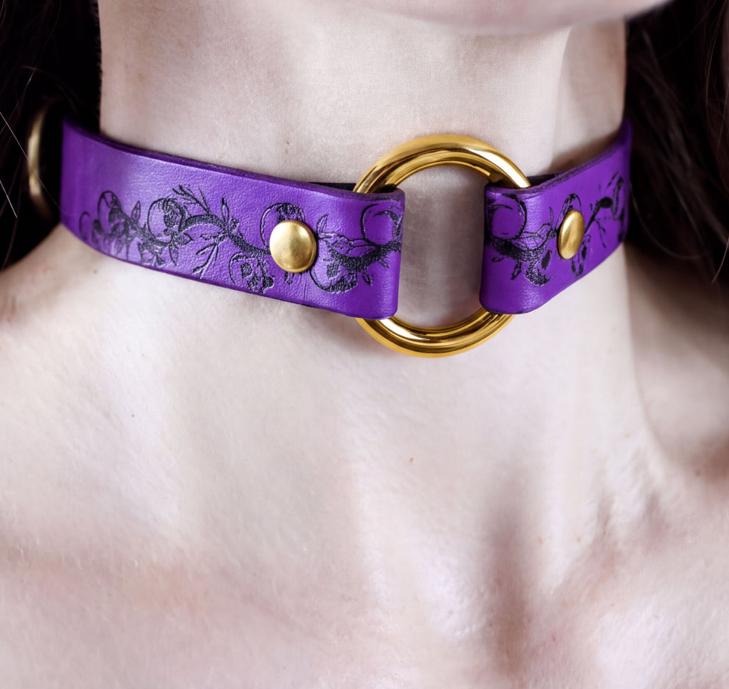 A close-up image of a person wearing a purple choker necklace with a gold ring and two gold buttons.