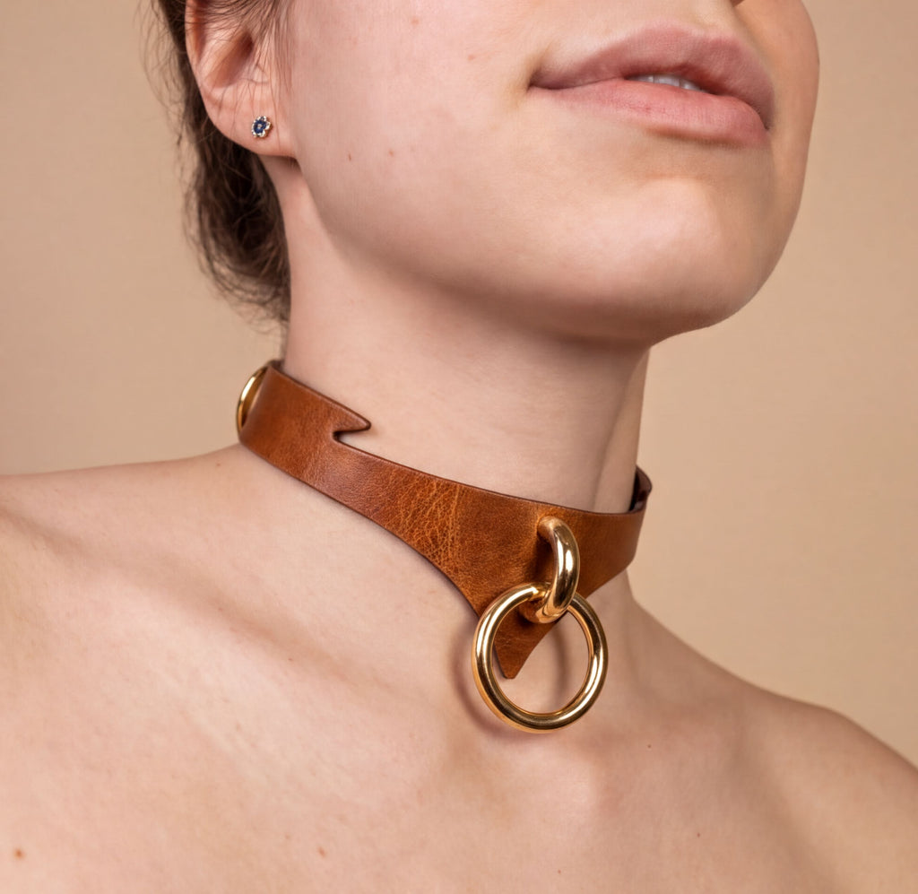 A close-up of a woman's neck and chest area, featuring a leather choker necklace with a gold-colored ring and a gold-colored buckle.